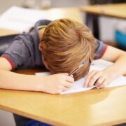 Student with blond hair rests head on desk while writing on paper with a blue pen in a classroom setting.
