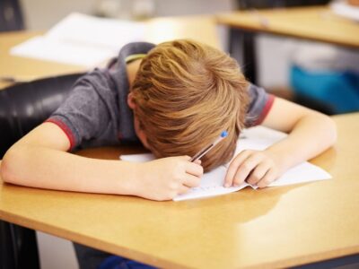 Student with blond hair rests head on desk while writing on paper with a blue pen in a classroom setting.