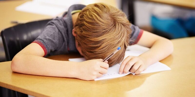 Student with blond hair rests head on desk while writing on paper with a blue pen in a classroom setting.