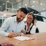 Man signs paperwork at a dealership desk, smiling woman beside him with a white car in the background on display.