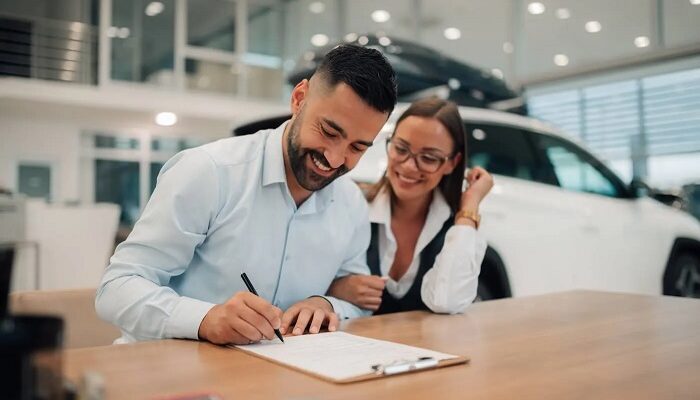 Man signs paperwork at a dealership desk, smiling woman beside him with a white car in the background on display.