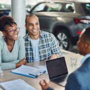 Smiling couple meeting with a car dealer at a dealership, reviewing documents with a laptop on the table and cars in the background,