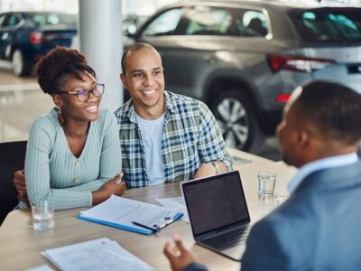 Smiling couple meeting with a car dealer at a dealership, reviewing documents with a laptop on the table and cars in the background,