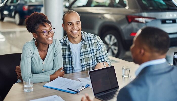Smiling couple meeting with a car dealer at a dealership, reviewing documents with a laptop on the table and cars in the background,