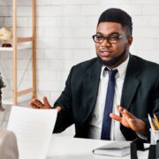 Professional man in a suit and glasses explains something to a colleague across a desk in a bright office setting with papers on the desk.
