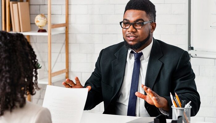 Professional man in a suit and glasses explains something to a colleague across a desk in a bright office setting with papers on the desk.