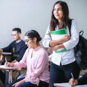 A classroom scene with a smiling student standing with a backpack and notebooks while peers take notes at their desks in the background.