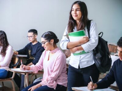 A classroom scene with a smiling student standing with a backpack and notebooks while peers take notes at their desks in the background.