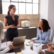 Woman presenting to a small team in a bright office; three colleagues listen around a table with a laptop and papers.
