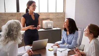 Woman presenting to a small team in a bright office; three colleagues listen around a table with a laptop and papers.