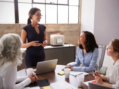 Woman presenting to a small team in a bright office; three colleagues listen around a table with a laptop and papers.