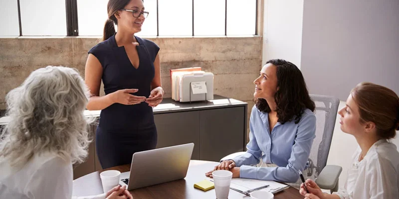 Woman presenting to a small team in a bright office; three colleagues listen around a table with a laptop and papers.