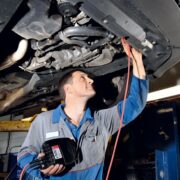 Auto mechanic in blue coveralls inspects the underside of a car on a lift, holding a diagnostic tool and red test lead connected to the vehicle.