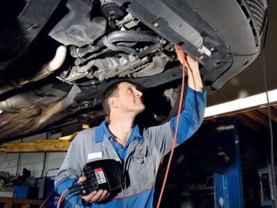 Auto mechanic in blue coveralls inspects the underside of a car on a lift, holding a diagnostic tool and red test lead connected to the vehicle.