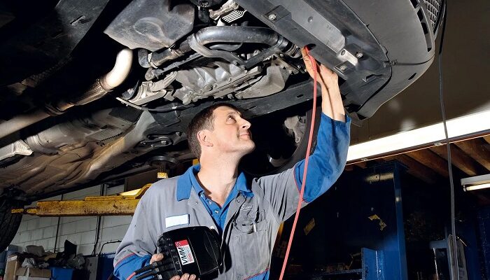 Auto mechanic in blue coveralls inspects the underside of a car on a lift, holding a diagnostic tool and red test lead connected to the vehicle.
